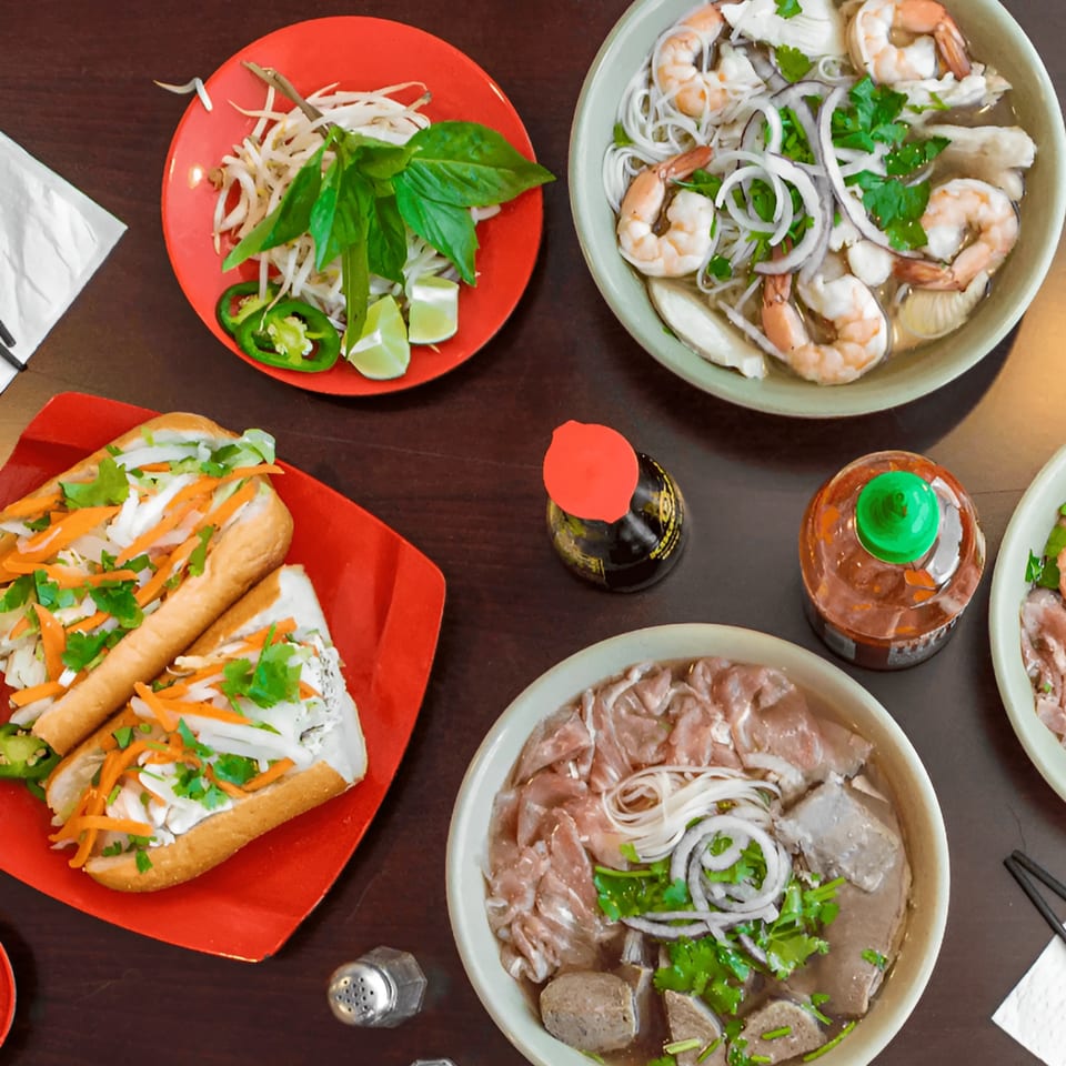Overhead view of Vietnamese pho with shrimp and beef, banh mi sandwiches, and fresh garnishes.